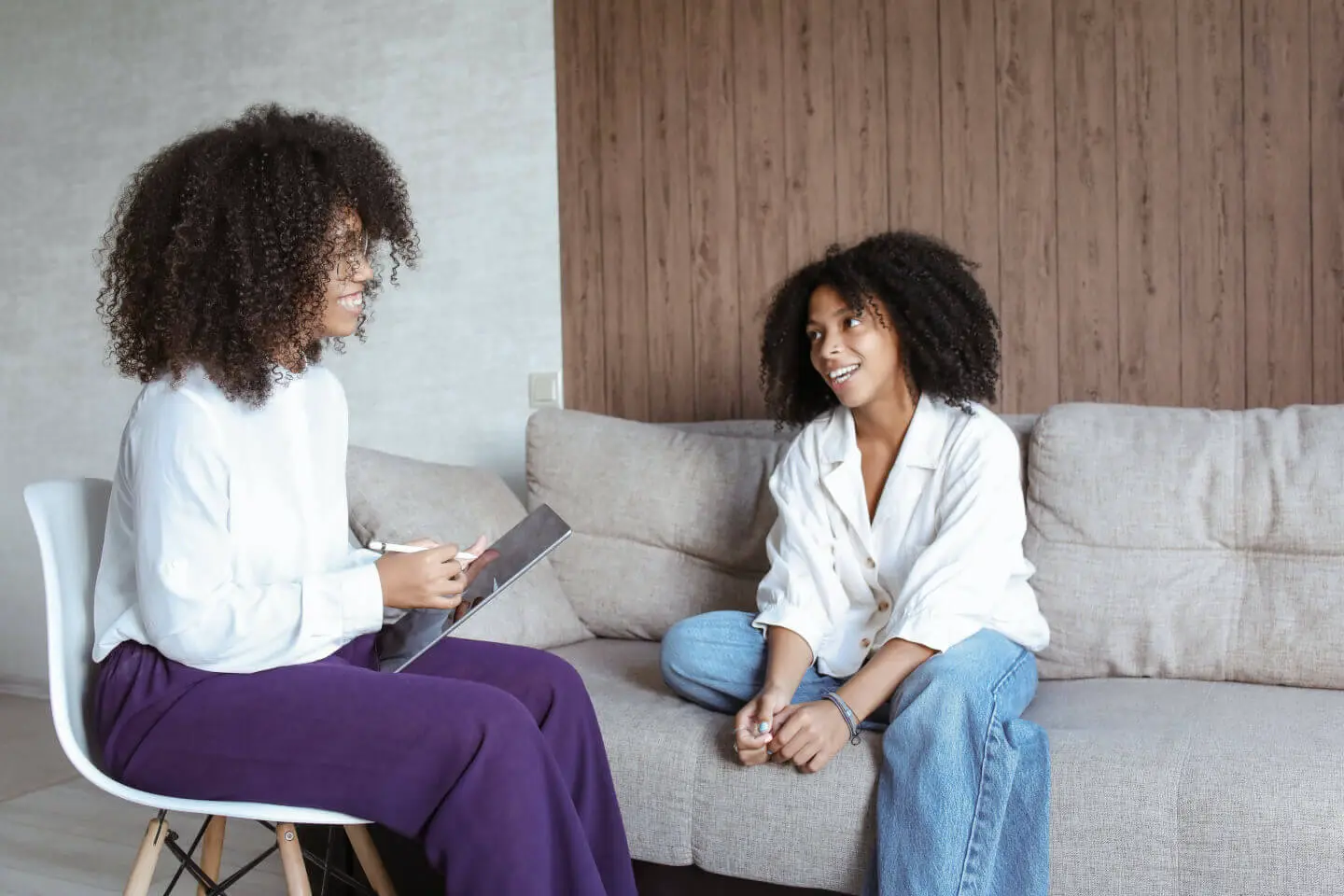 A woman sitting on a couch speaking to a therapist who is sitting in a chair across from her.