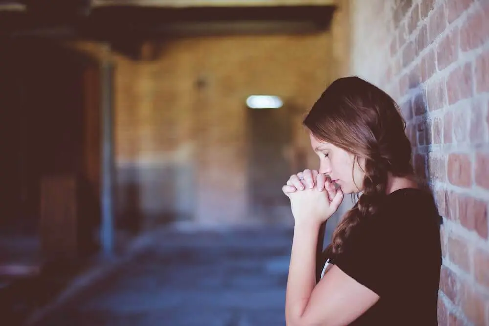 A lady leaning her back against a brick wall closes her eyes and raises her hands in a prayer position under her face.