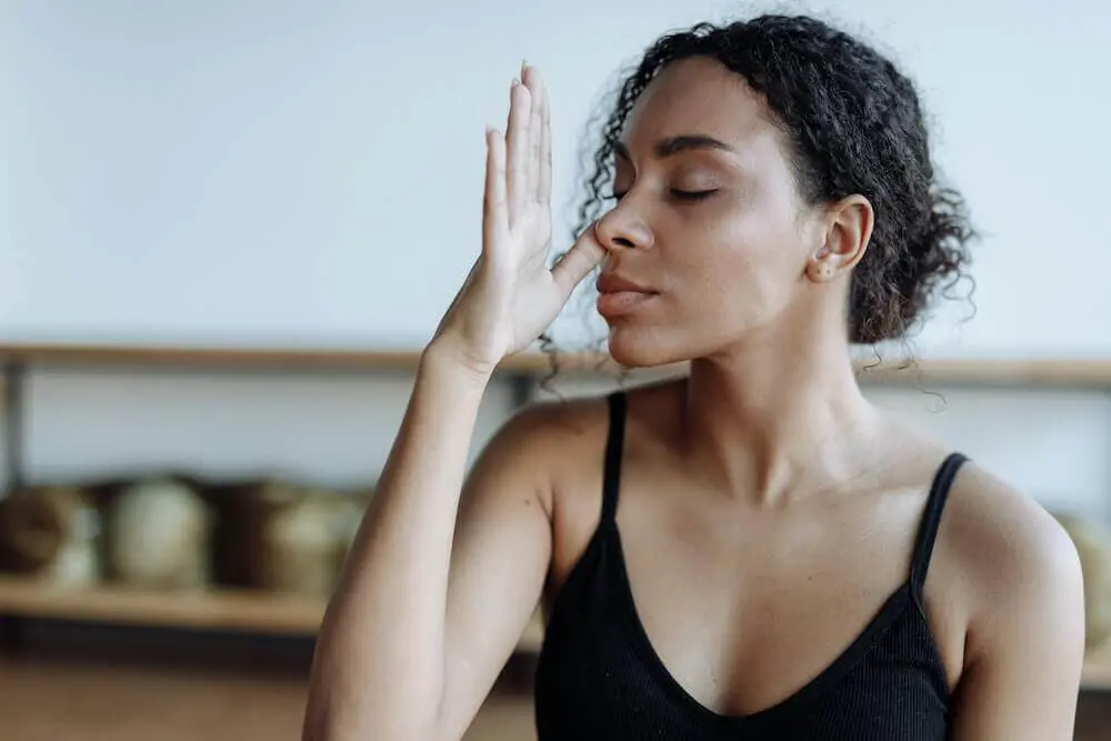 Woman in black tank top sitting with eyes closed and thumb to their nose doing a breathing exercise