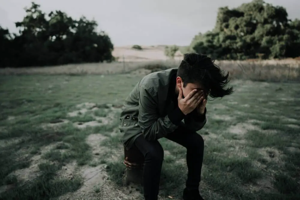 A man sits with his hands in his face in the middle of a field. 