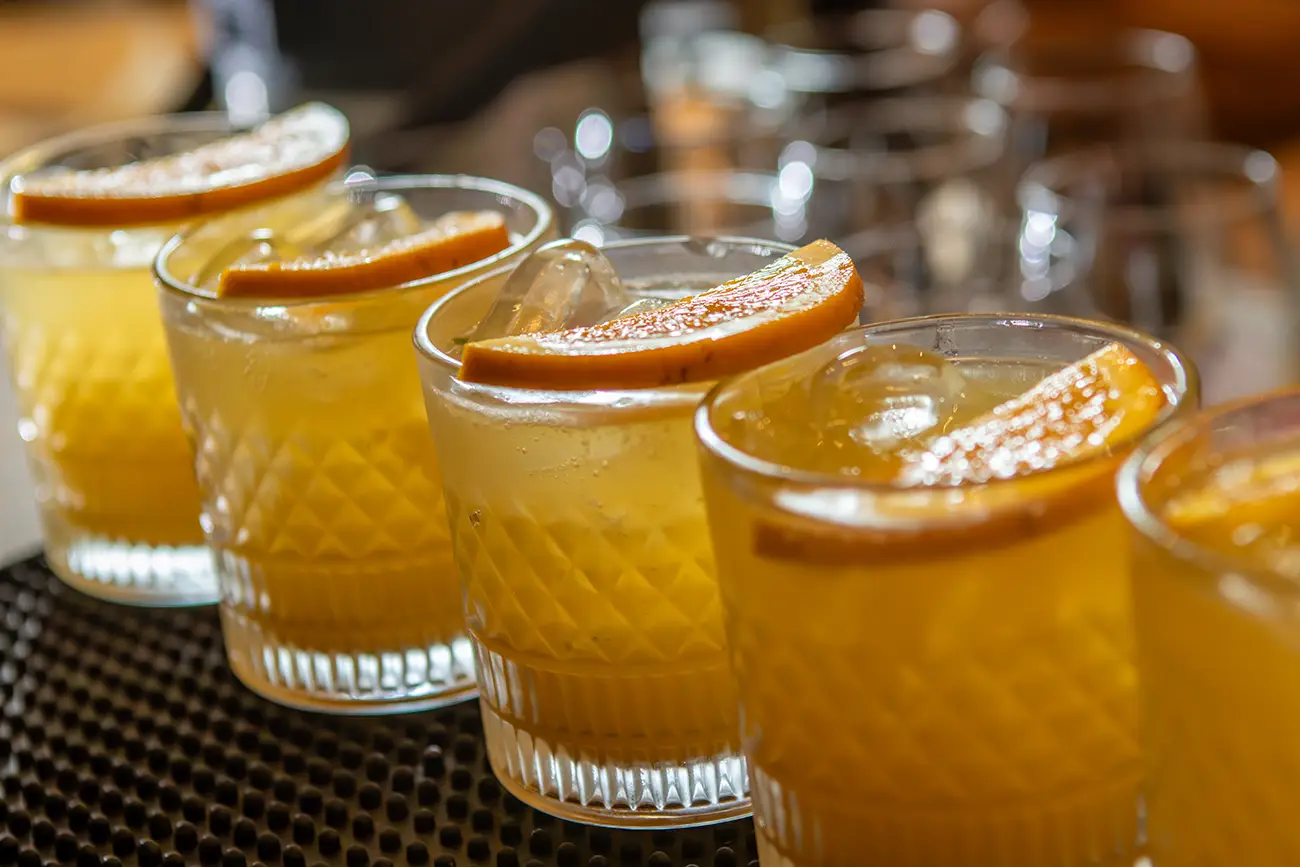 Cinco copos com coquetel amarelo, gelo e fatia de laranja em cima, alinhados sobre uma superfície preta texturizada.