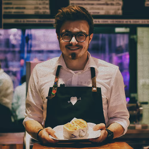Smiling man with glasses wearing a black apron holding a plate with two tortilla wraps.
