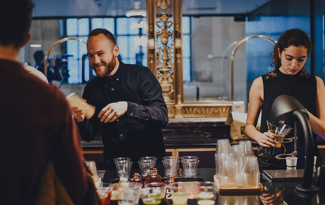 Barista shaking a cocktail shaker and smiling while another barista pours coffee from a Chemex at a cafe counter.