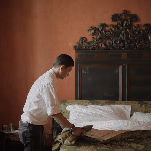 A man in a white shirt making a bed in a room with ornate furniture and warm brown walls.