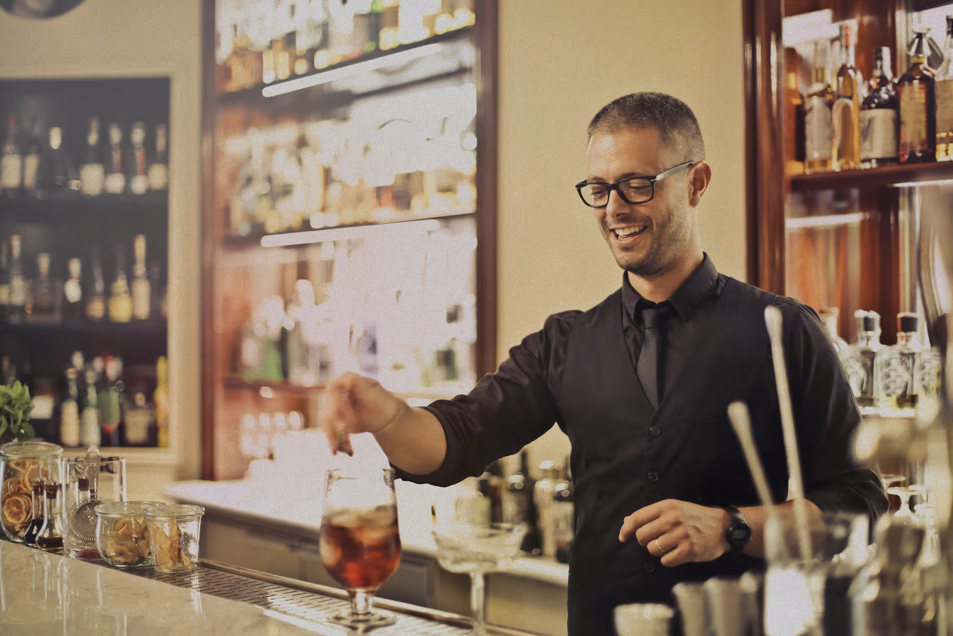 Smiling bartender in black shirt and tie preparing a cocktail behind a bar counter with jars of ingredients.