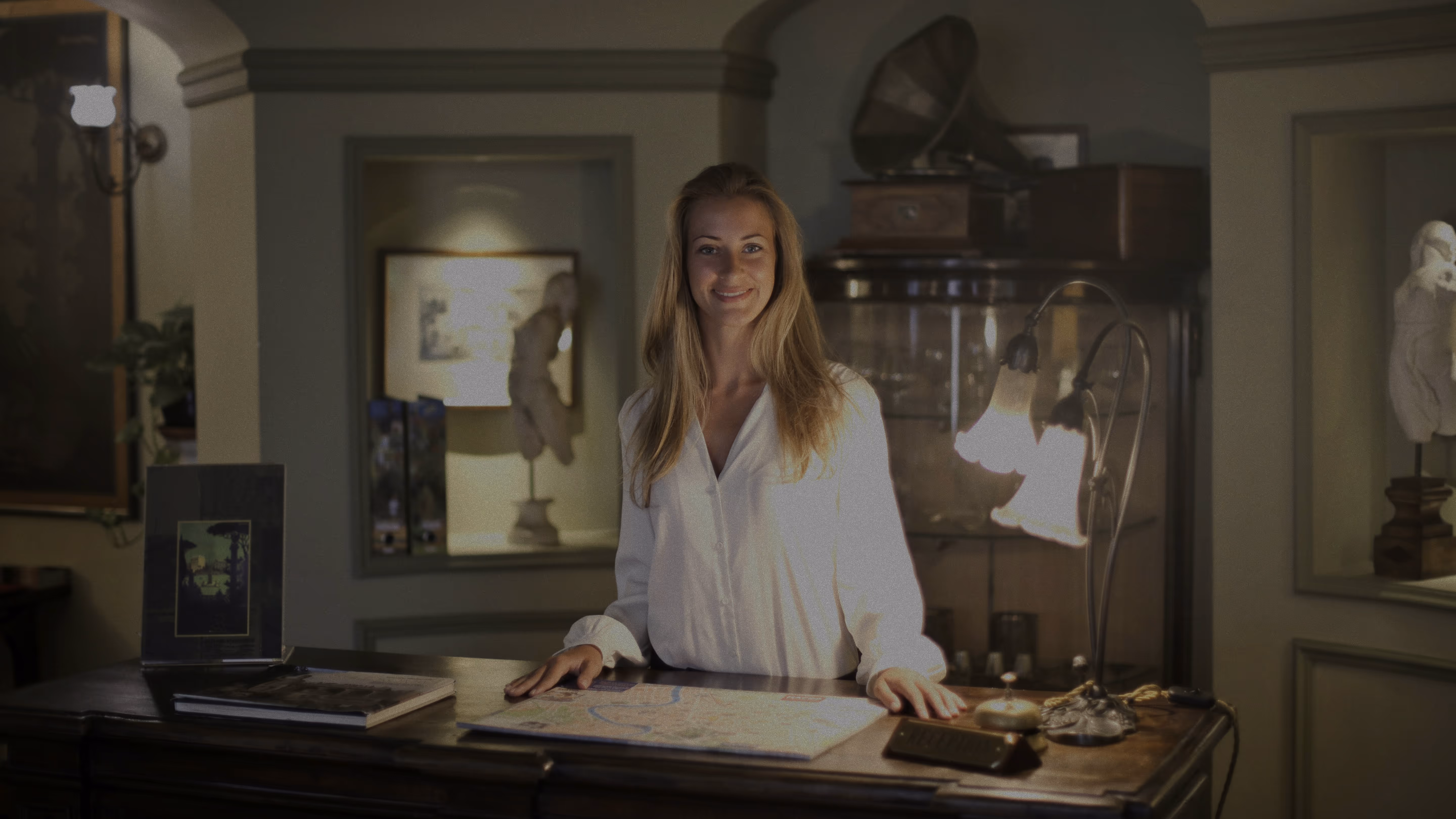 Smiling woman with long blonde hair standing behind a wooden reception desk in a warmly lit vintage room.