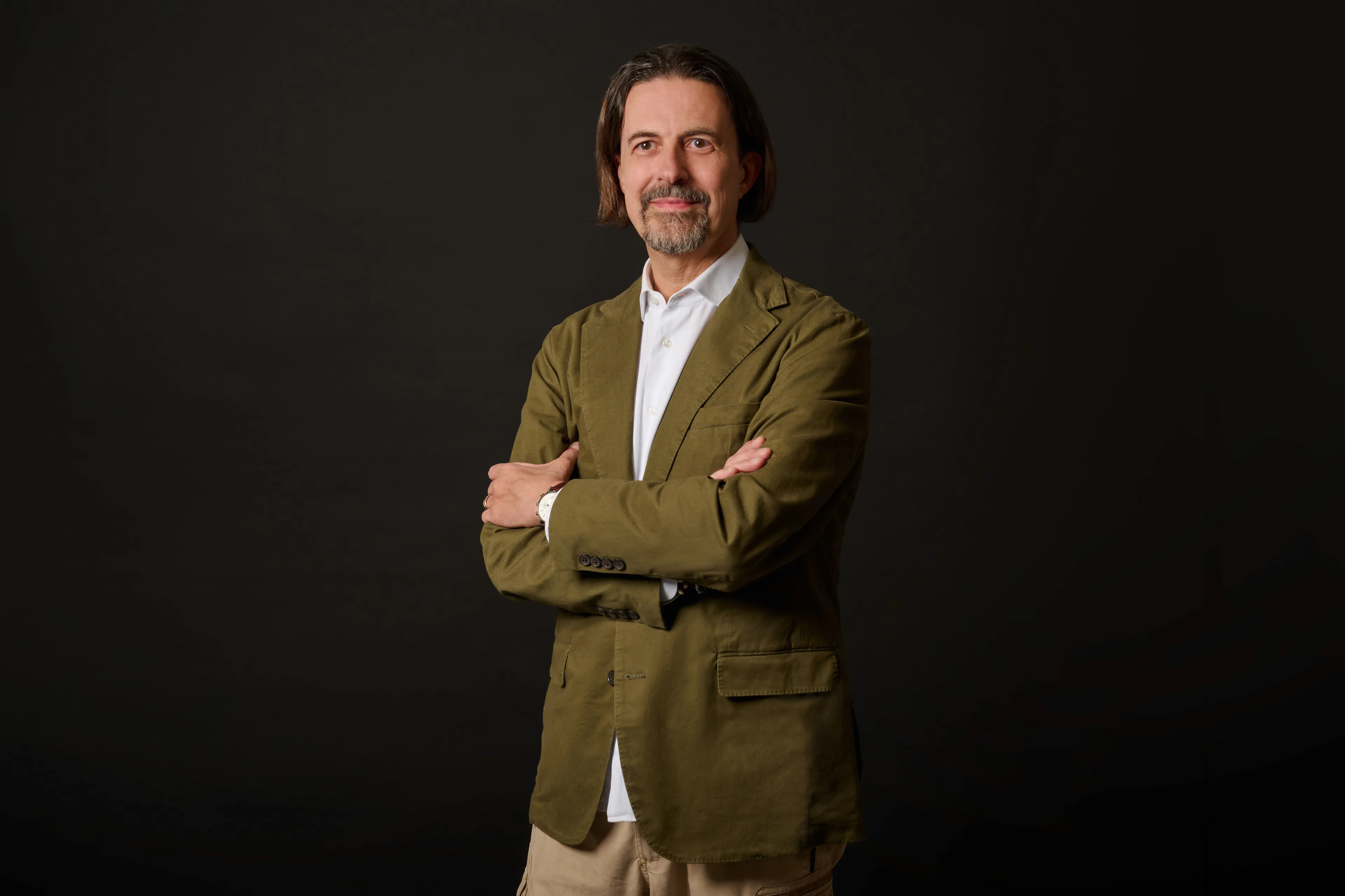 Man with medium-length hair and beard wearing an olive green jacket and white shirt standing with arms crossed.