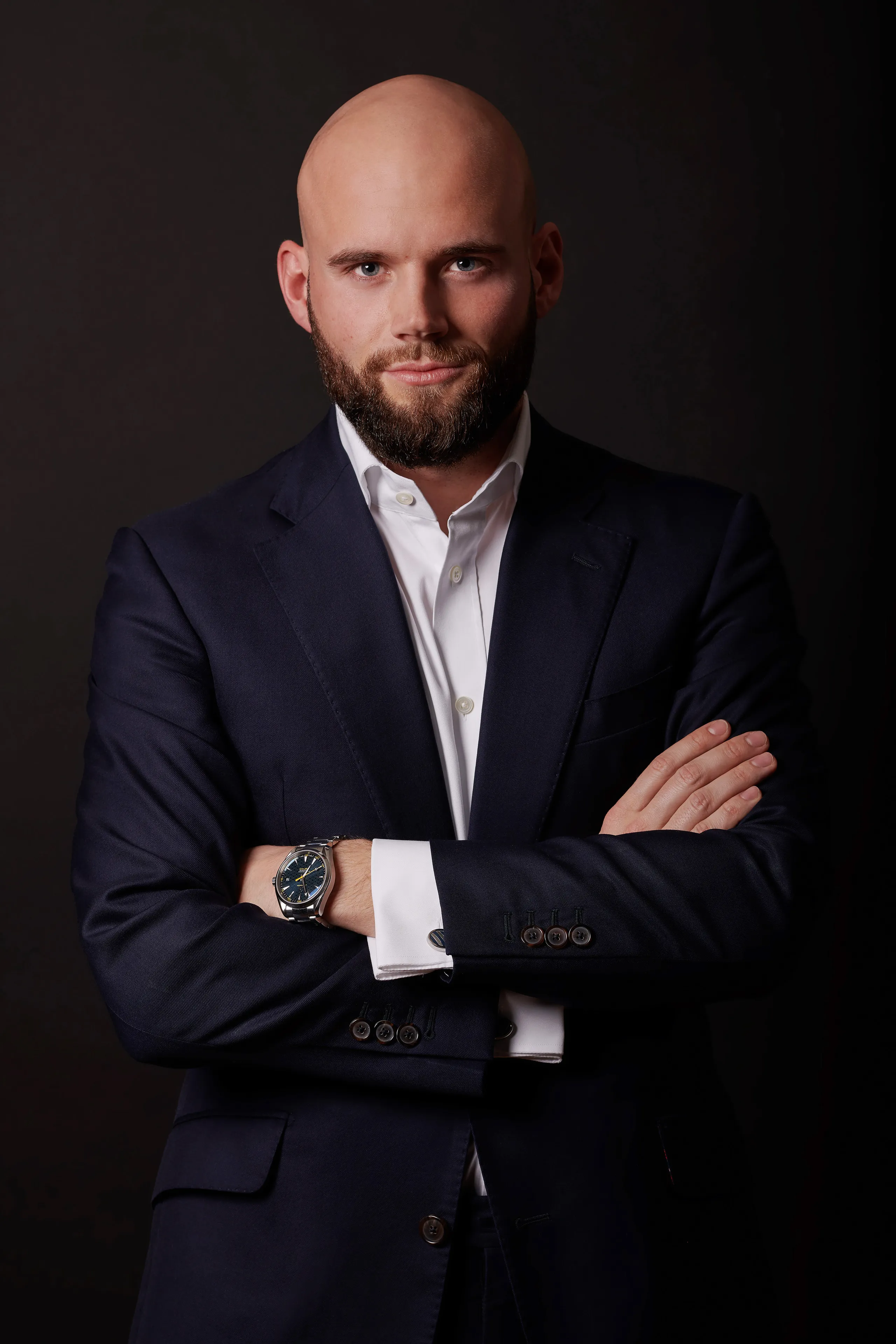 Confident bald man with beard in white shirt and navy suit, arms crossed against dark background.
