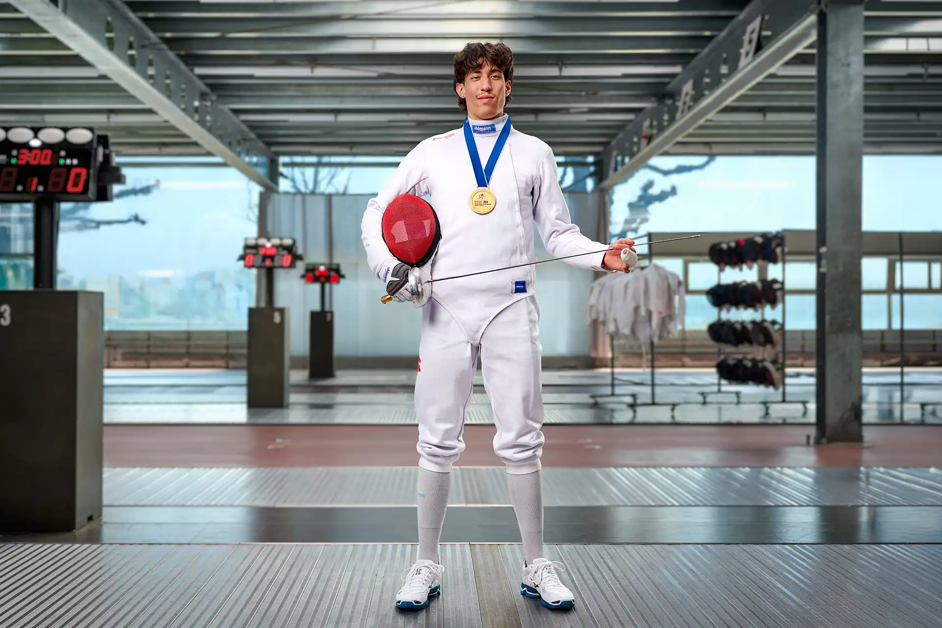 Fencer in white gear holding mask and foil, wearing gold medal, standing in a fencing hall.