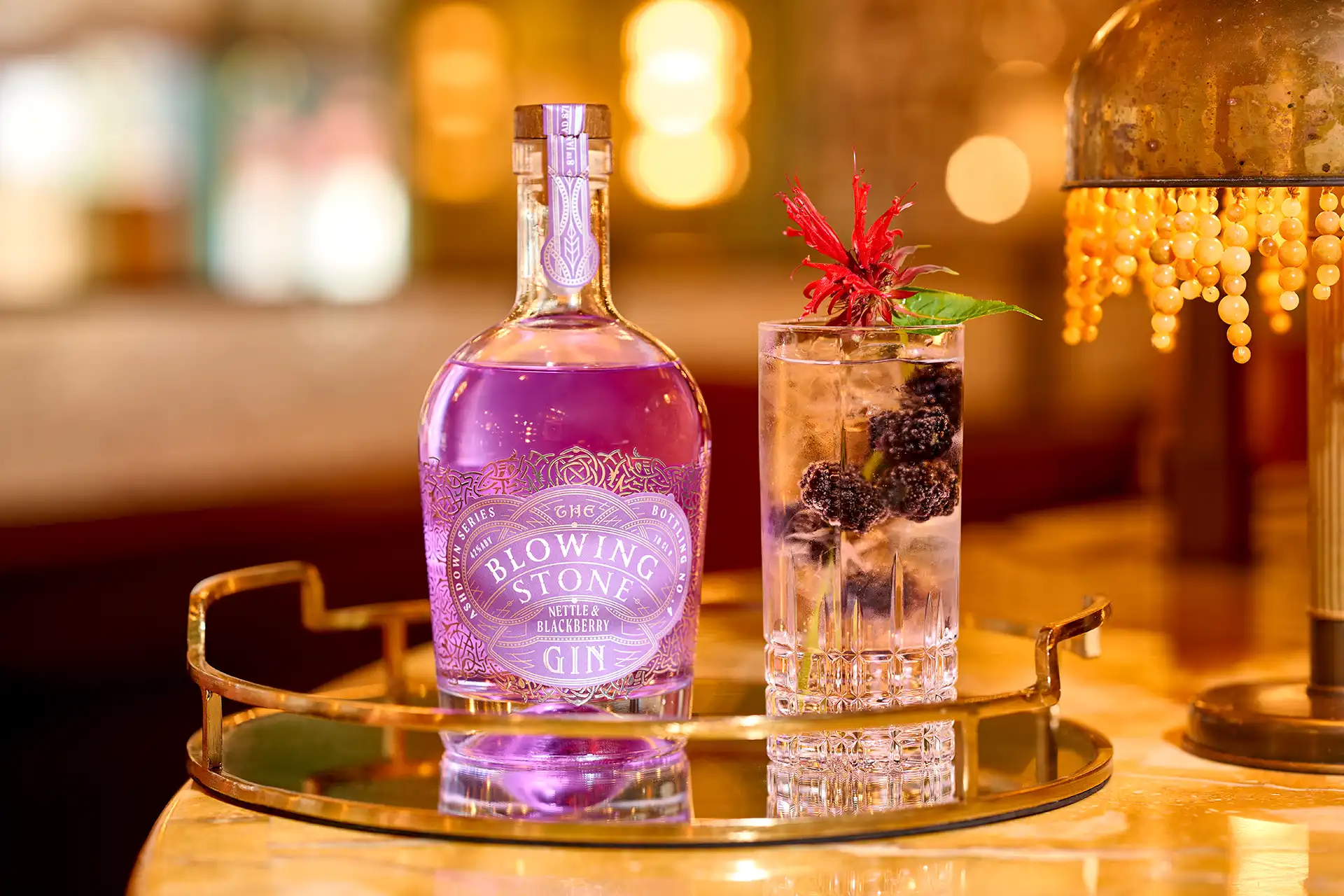 Purple bottle of Blowing Stone gin next to a glass with blackberries and a red flower garnish.