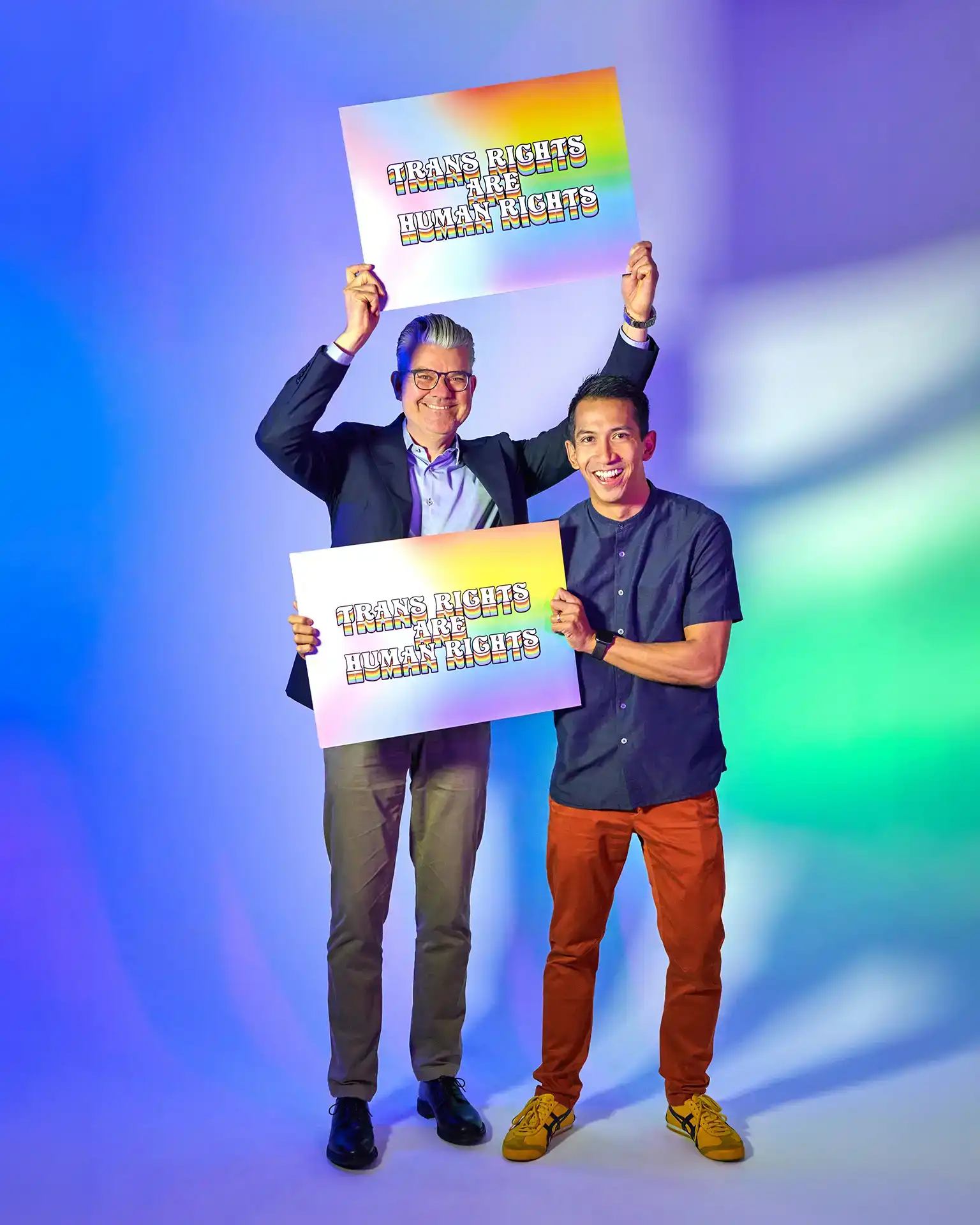 Two men smiling and holding rainbow signs reading 'Trans rights are human rights' against a colorful background.