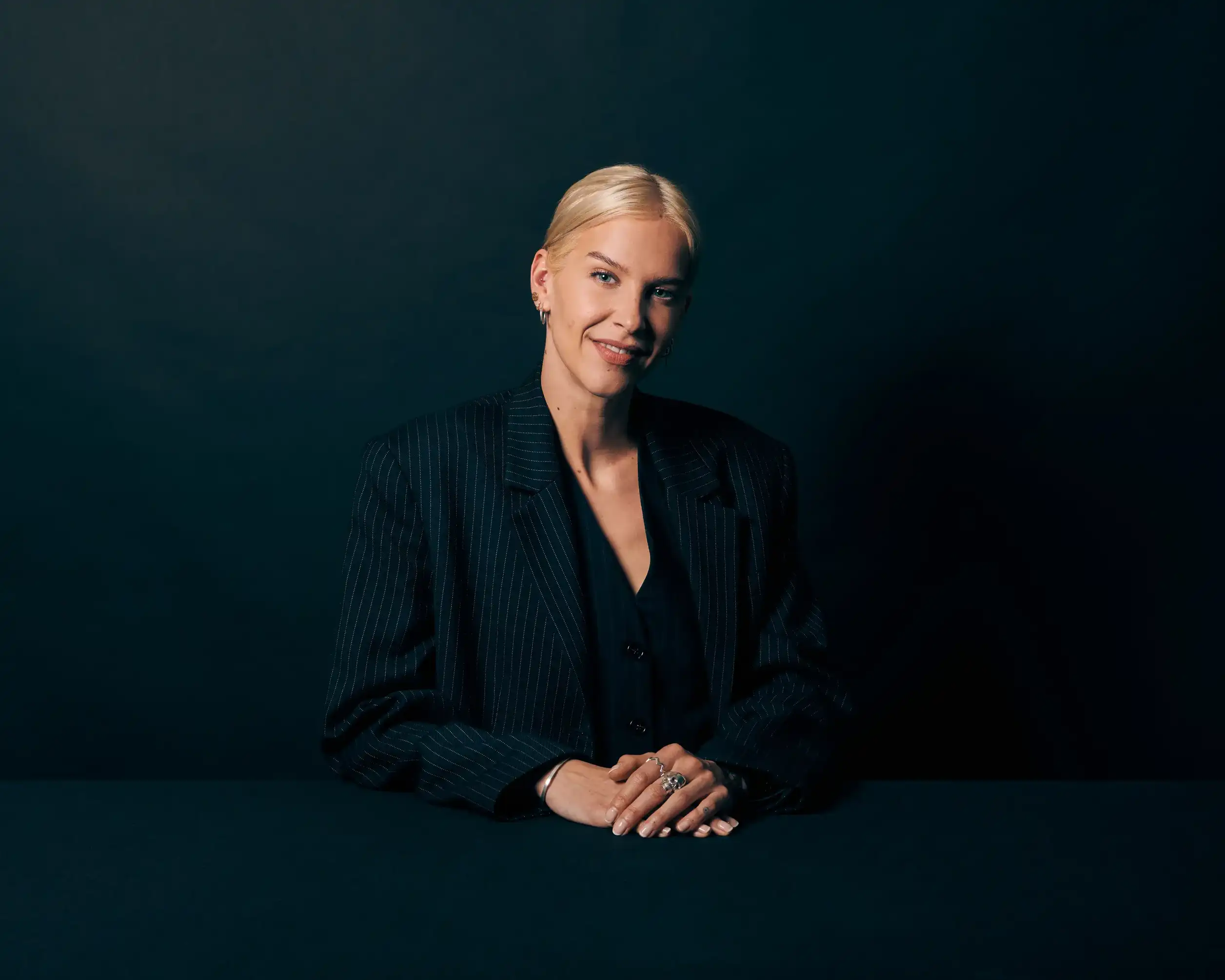 Smiling blonde woman wearing a dark pinstripe blazer, seated with hands crossed on a dark table.