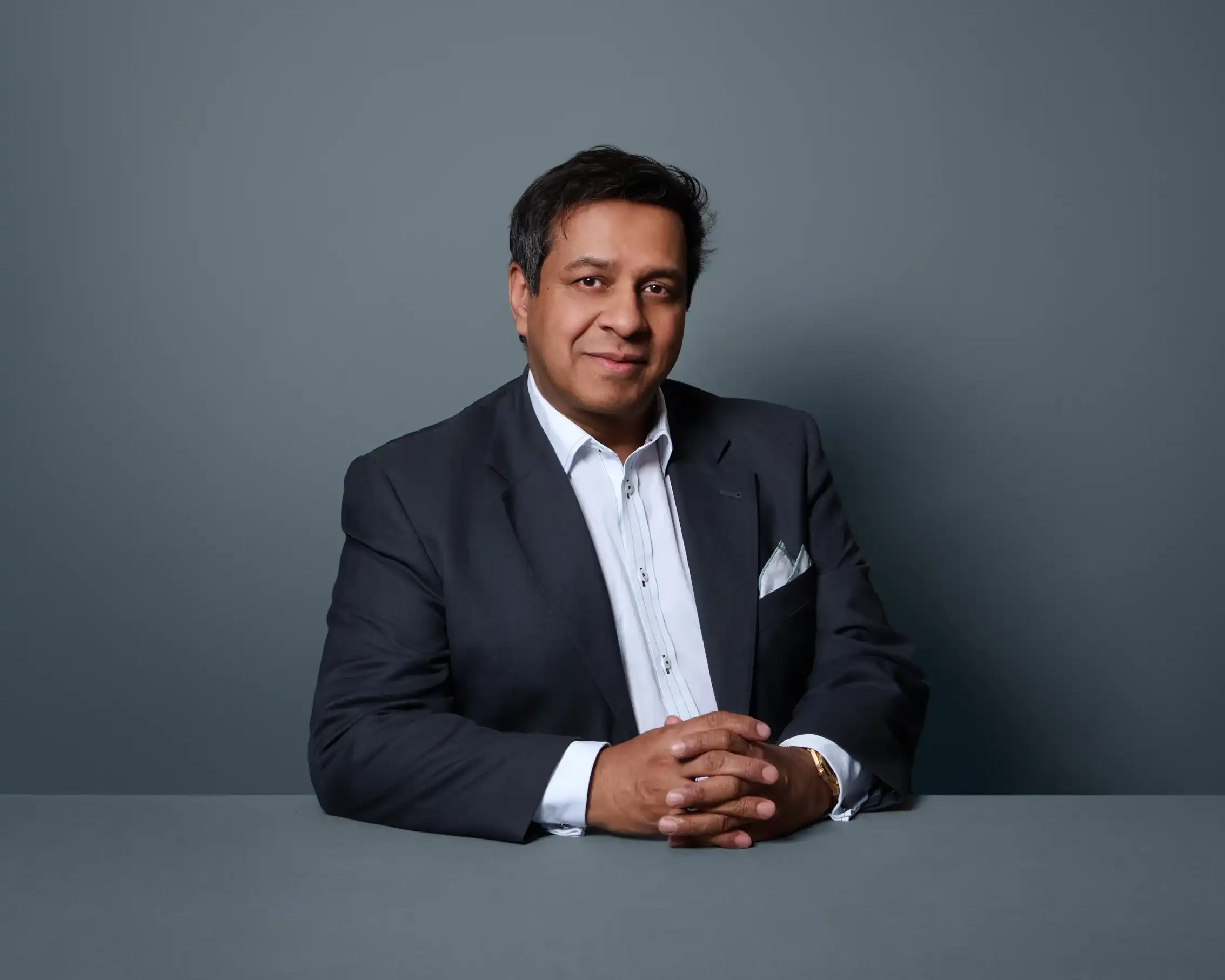 Man in dark suit and white shirt sitting with hands clasped on gray table against gray background.
