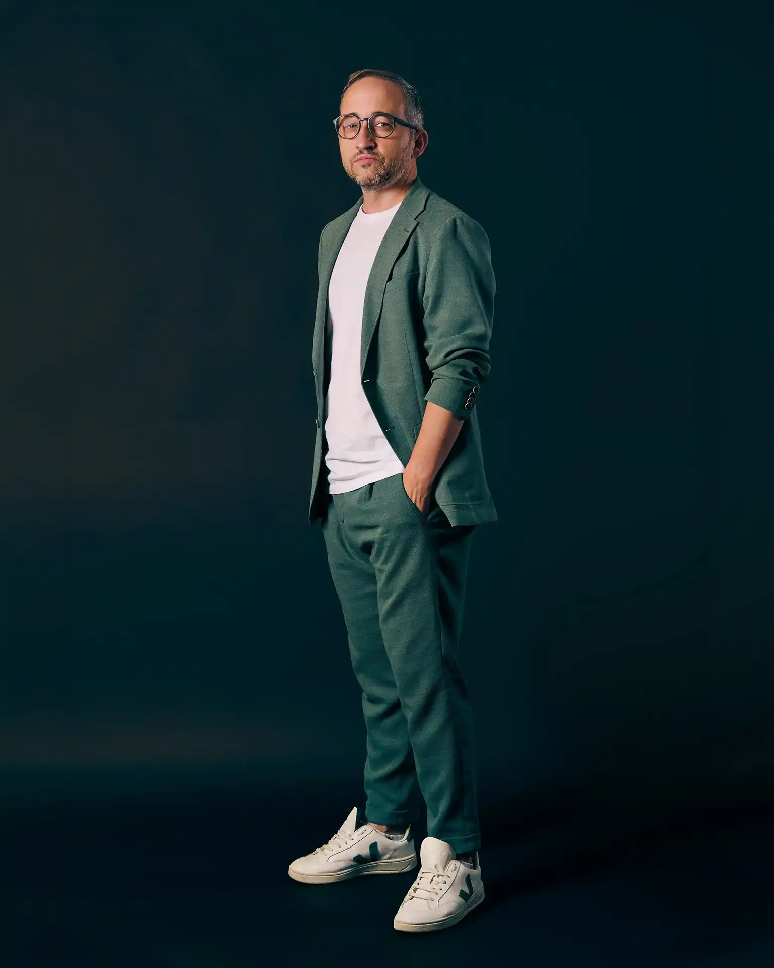 Man with glasses wearing green suit, white t-shirt, and white sneakers, standing with one hand in pocket.