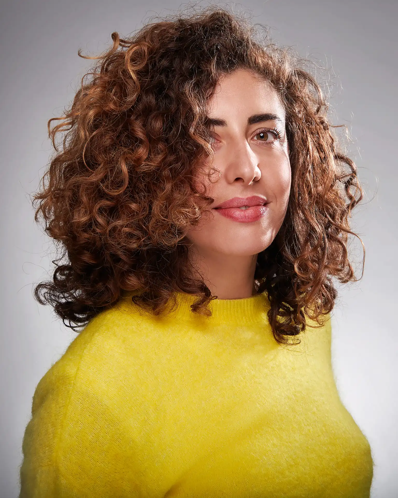 Woman with voluminous curly hair wearing a bright yellow fuzzy sweater against a gray background.