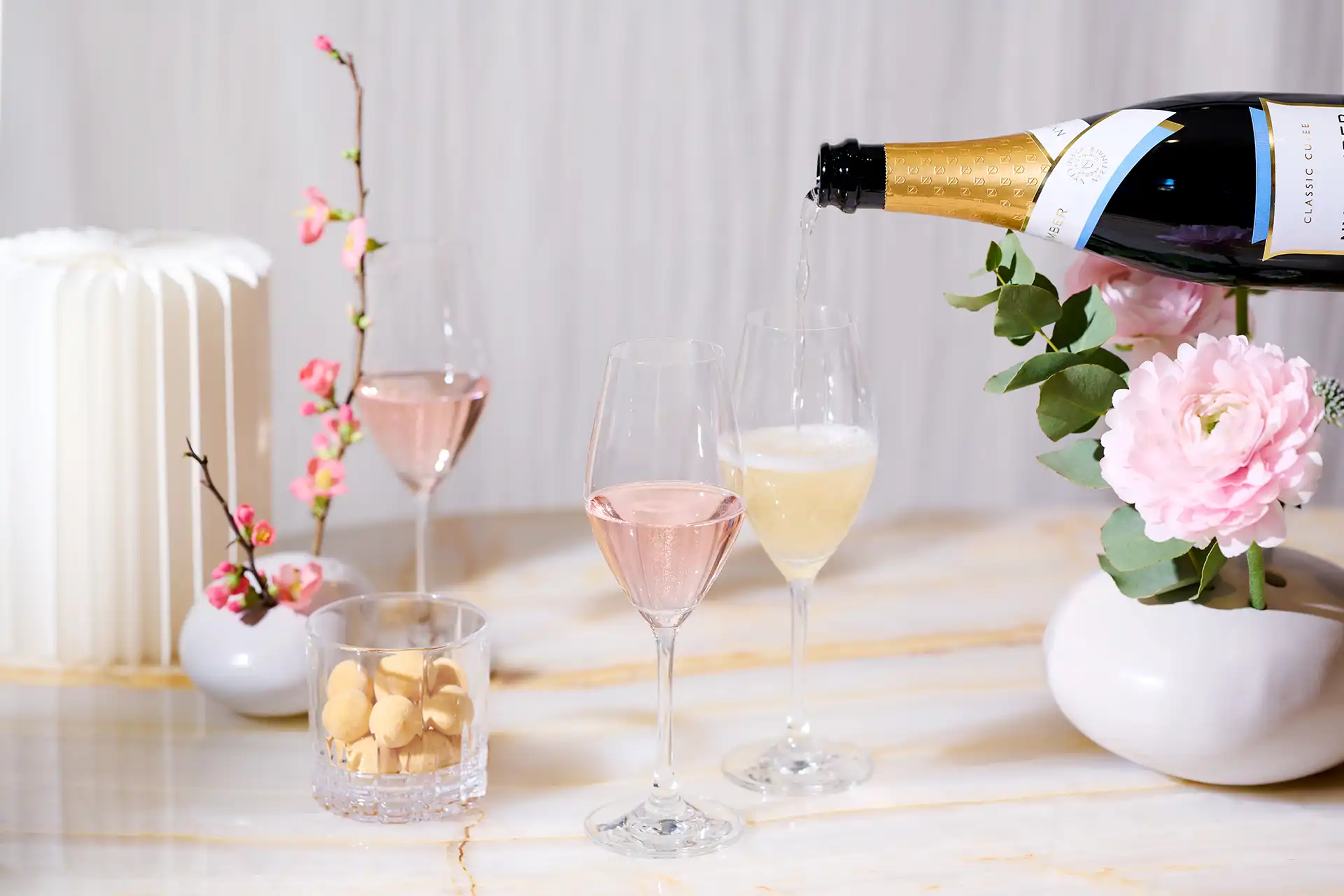 Glasses of rosé and sparkling wine being poured, with pink flowers and a white candle on a marble table.