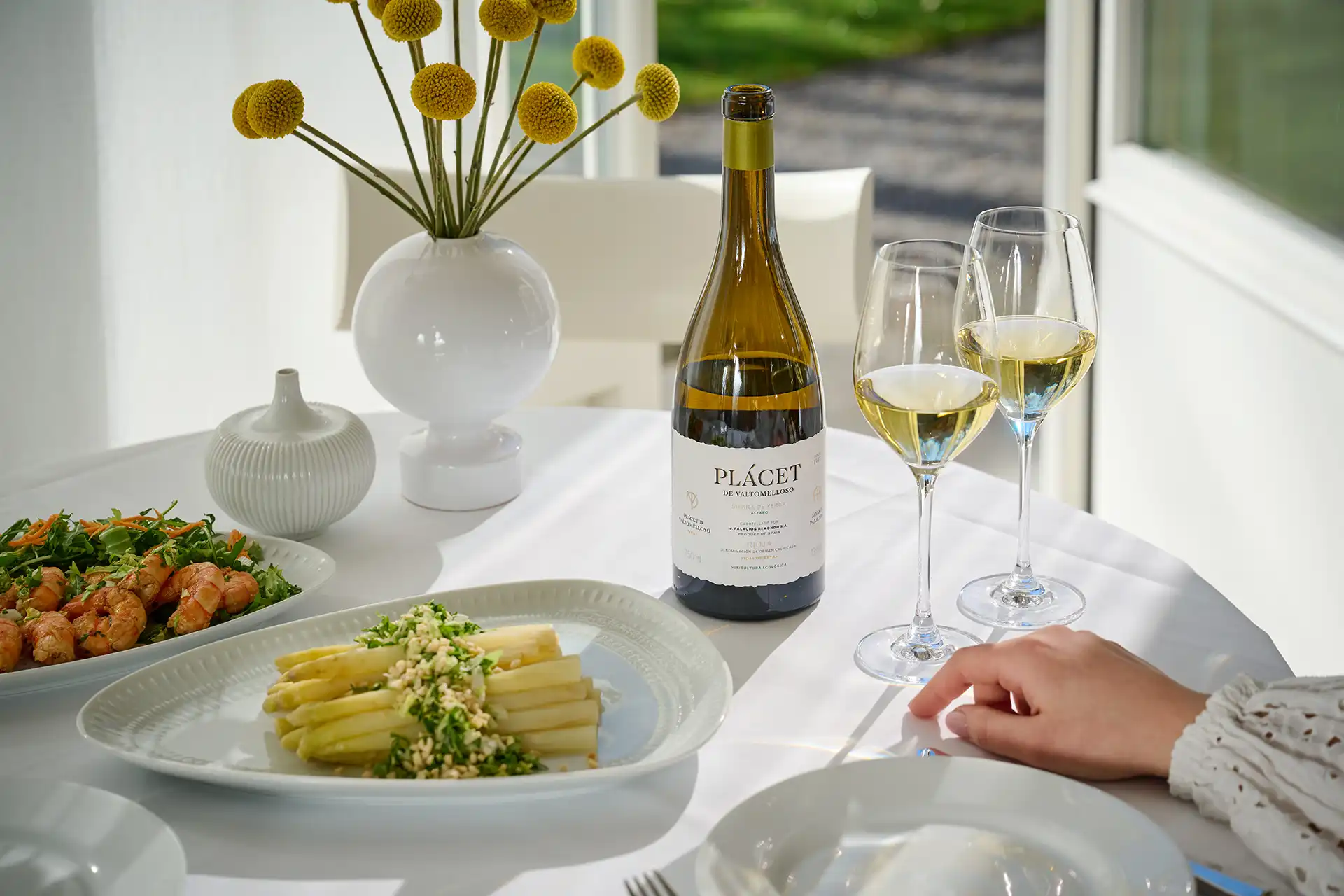 Table with white wine bottle, two filled glasses, plates of shrimp and asparagus, and a white vase with yellow flowers.