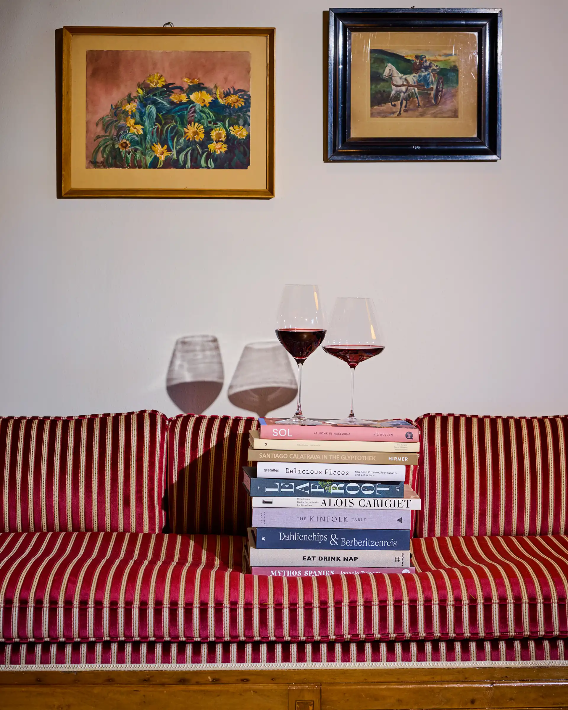Two wine glasses on a stack of books on a red-striped sofa below two framed paintings on a wall.