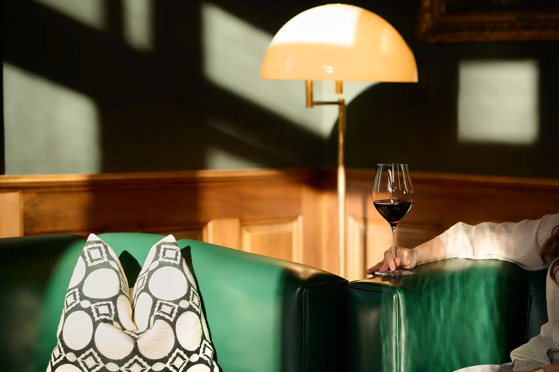 Green leather couch with patterned pillows and a hand holding a glass of red wine near a lit lamp.