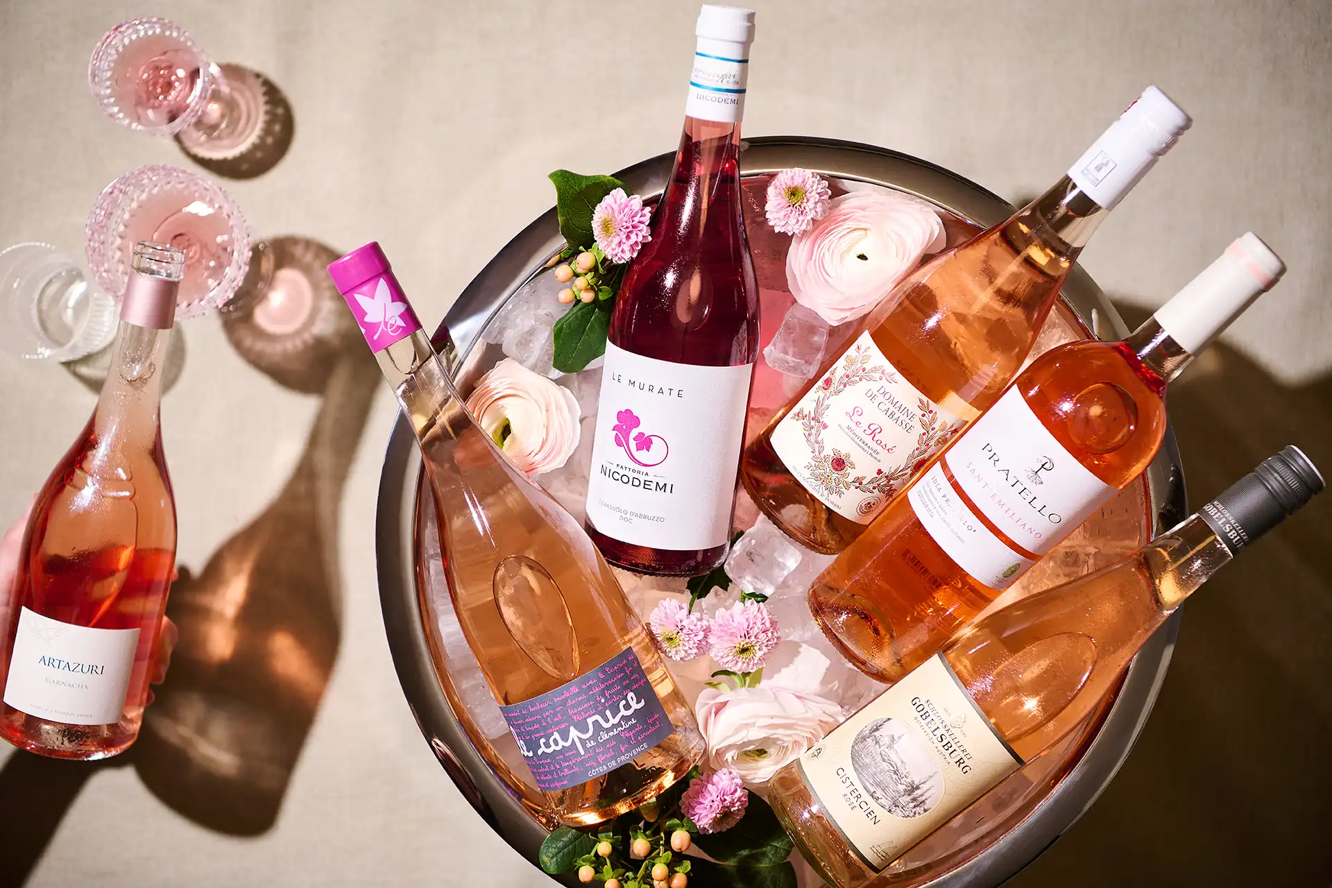 Ice bucket with six rosé wine bottles and pink flowers, one bottle being held above table.