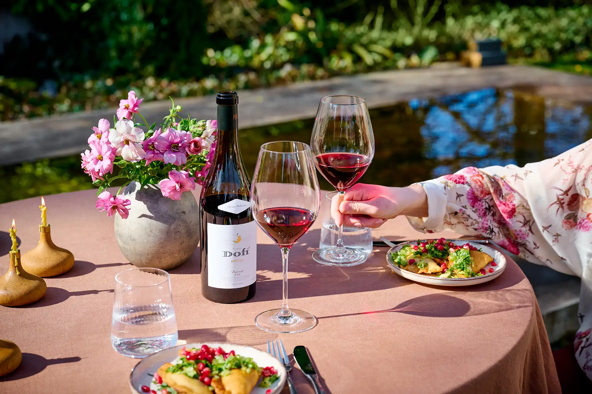 Outdoor table with wine bottle, two glasses of red wine, flowers, candles, water, and plates of food.