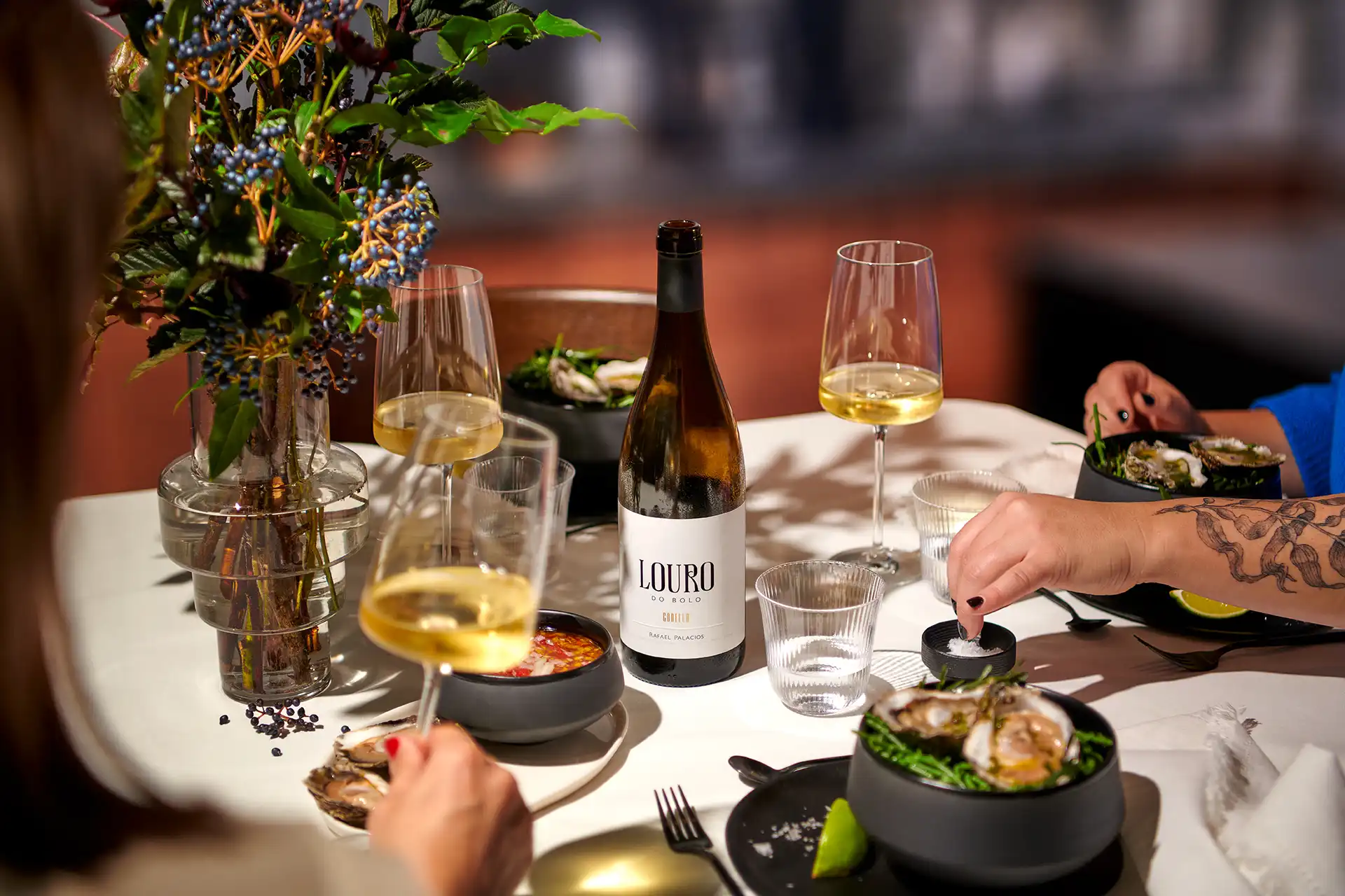 Two people dining with white wine, oysters, lime, and a bottle of Louro do Bolo on a table.