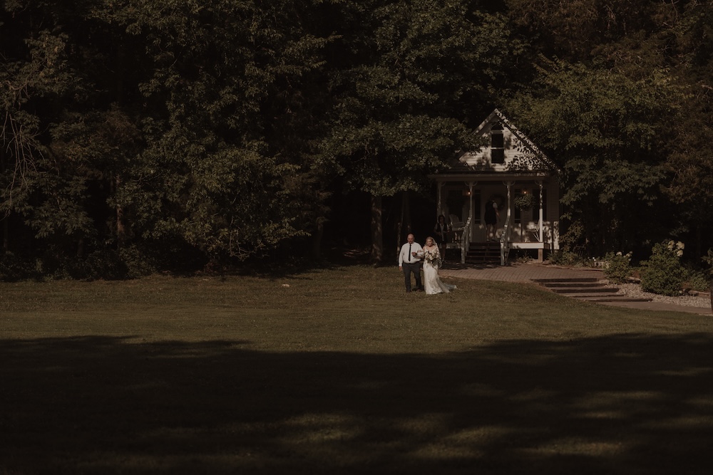 The bride and her father cross the lawn to the wedding ceremony with the bride's cottage in the background at The Barn at Cedar Grove between Columbia & Greensburg in Kentuckya