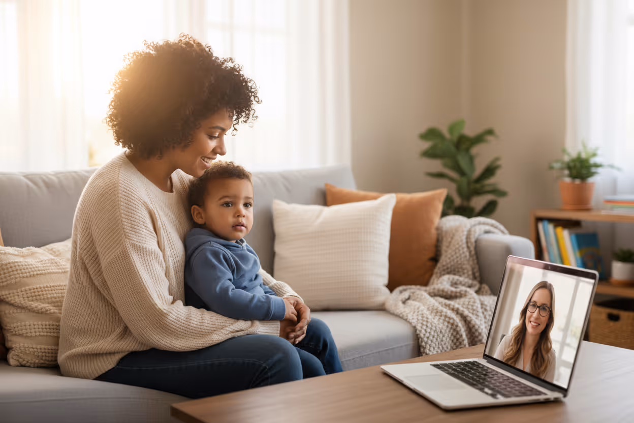 A mother and her young son sit on a couch having a video ABA therapy session with a smiling clinician on a laptop.