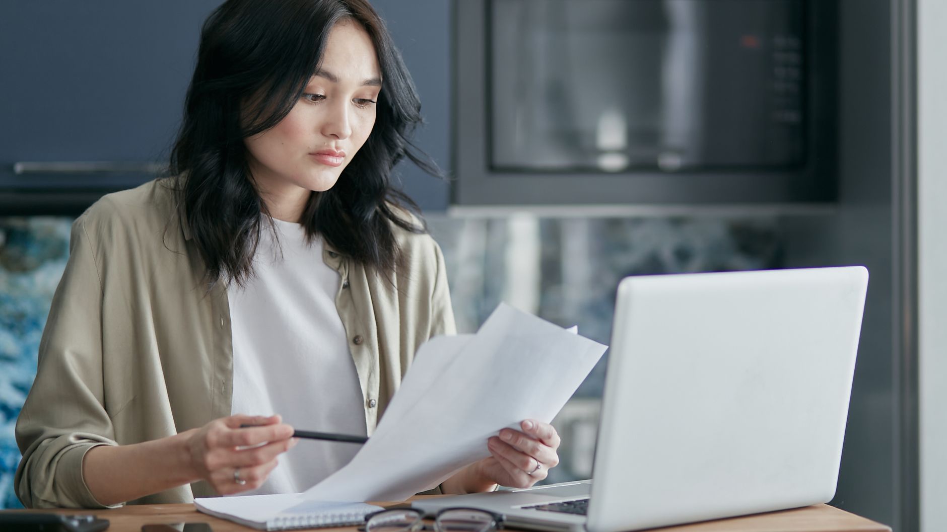 A parent reviewing printed paperwork beside an open laptop at a kitchen counter