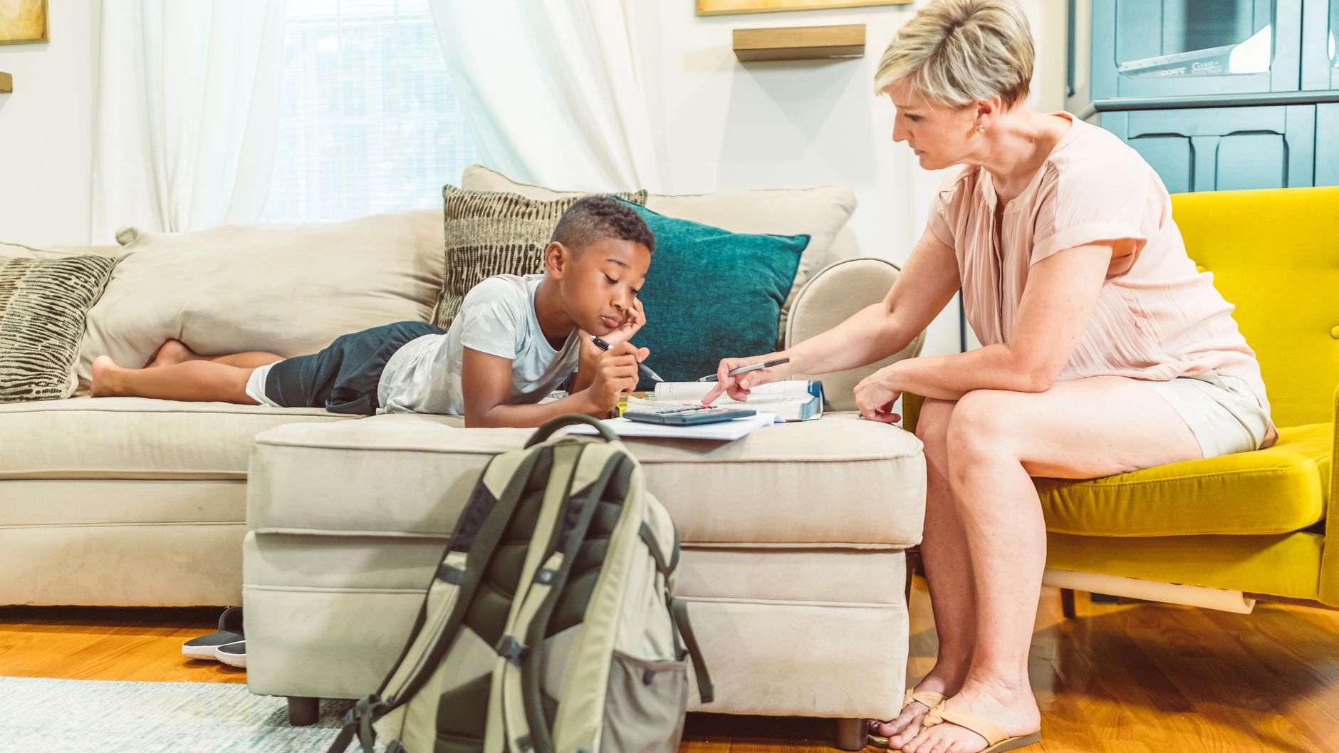 A school-age boy working on papers on an ottoman while a parent points at the page, a school backpack in the foreground