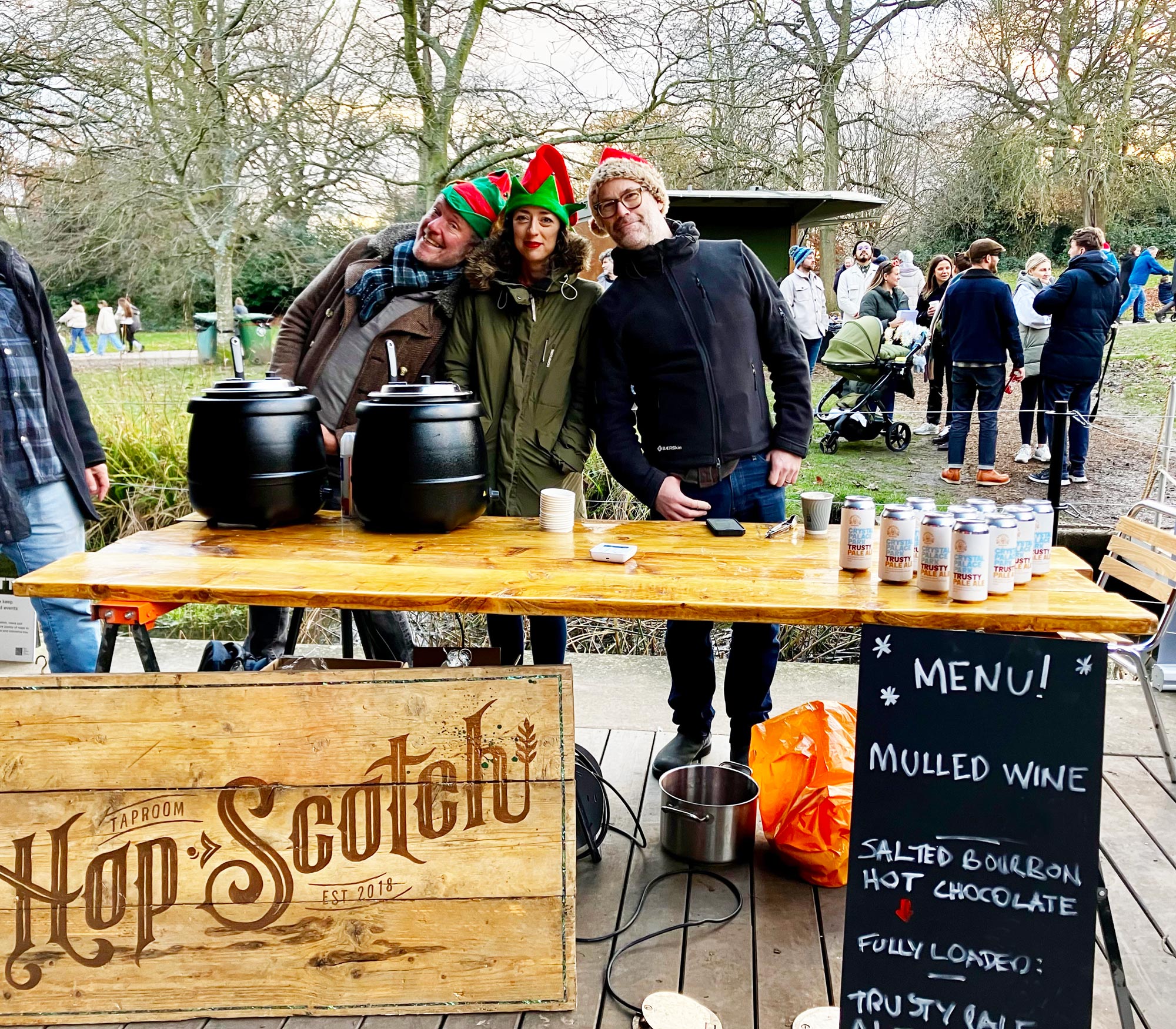 Three people wearing christmas themed hats stand being a wooden table with two black insulated heating pots and a wooden sign with the words HopScotch written