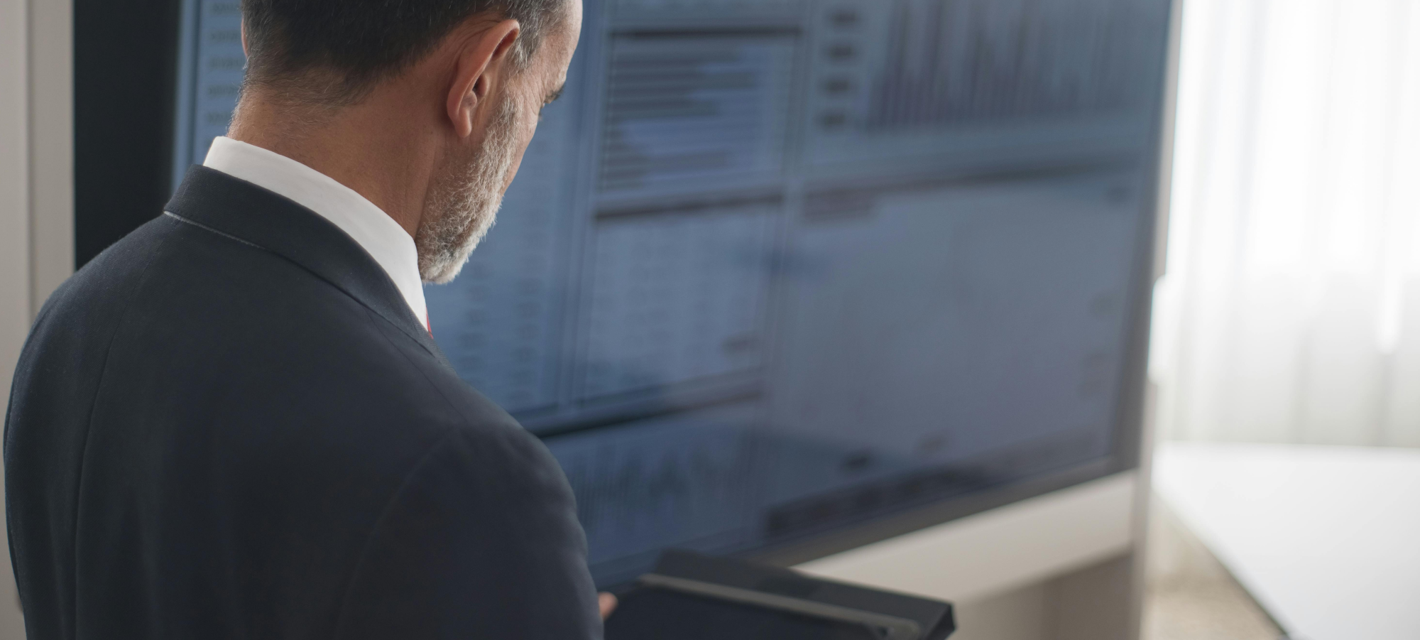 Ein Mann im Anzug betrachtet ein Tablet, während er vor einem großen Monitor steht, der verschwommen eine komplexe Datendarstellung und Diagramme zeigt. Die Szene vermittelt ein professionelles Umfeld, möglicherweise im Bereich Datenanalyse oder Business Intelligence.