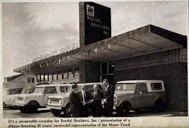 Black and white photo of three men standing in front of a building with International Trucks signage and two vintage trucks parked nearby.