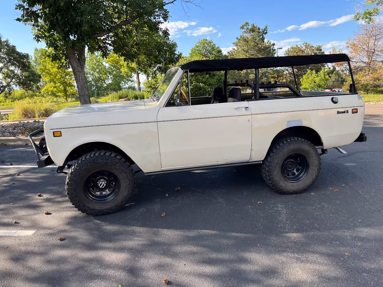 White vintage Scout II off-road vehicle with large black tires parked on an asphalt surface under trees.