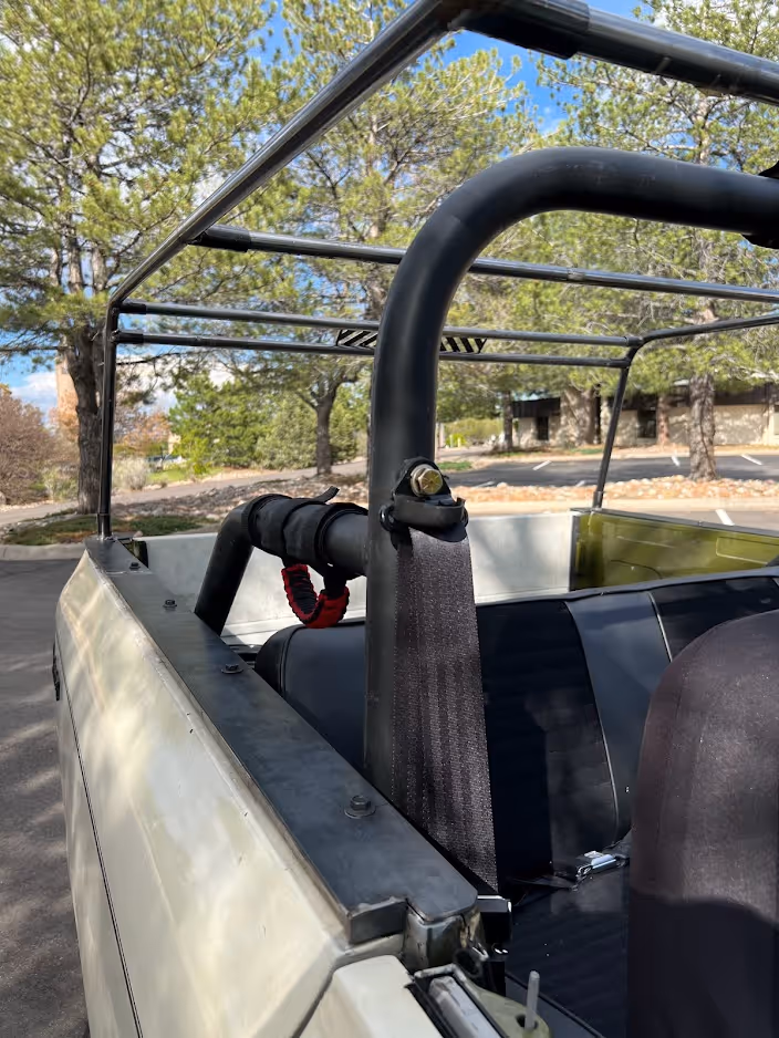 Interior view of a beige open-top vehicle with a black roll cage and black seatbelt, parked outdoors near trees.