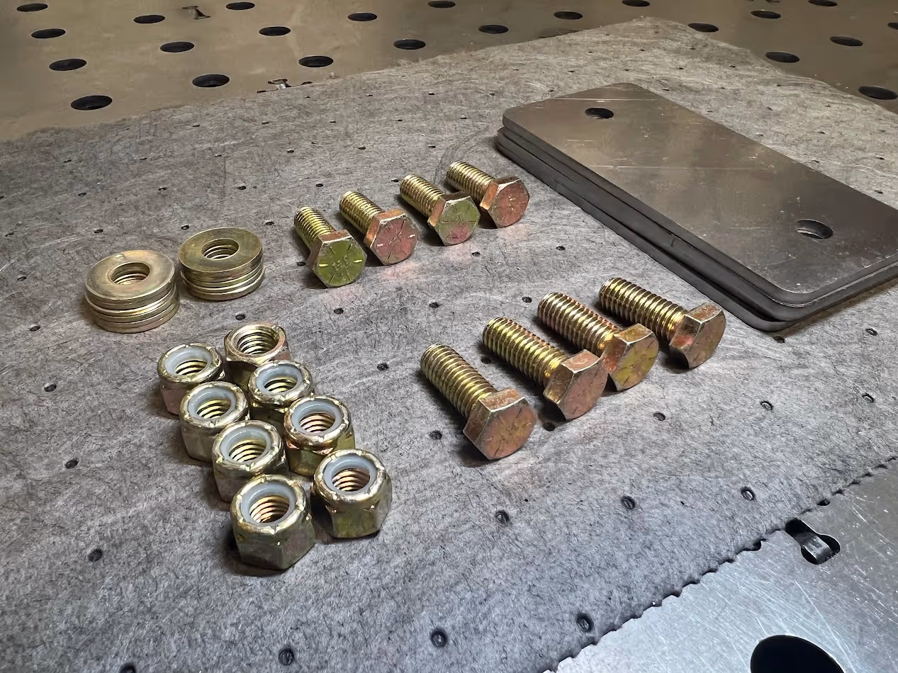 Metal bolts, nuts, washers, and rectangular metal plates laid out on a gray felt surface on a perforated metal workbench.