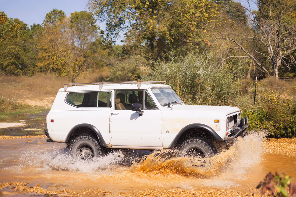 White off-road vehicle driving through a shallow muddy river with water splashing around the tires surrounded by green trees.
