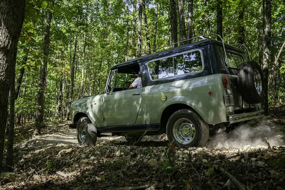 White off-road vehicle driving through a shallow muddy river with water splashing around the tires surrounded by green trees.