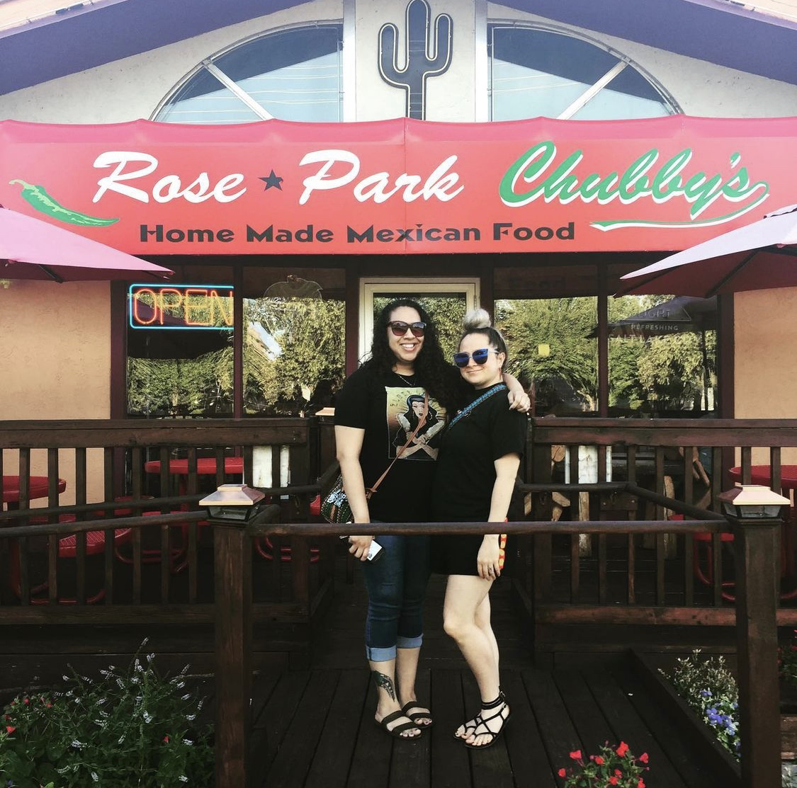 Molly and her friend stand on the deck in front of the Rose Park Chubby's restaurant. Greenery reflects in the restaurant's windows, a large red banner showing "Rose Park Chubby's" in green and white writing with decorative jalapeno hangs overhead.