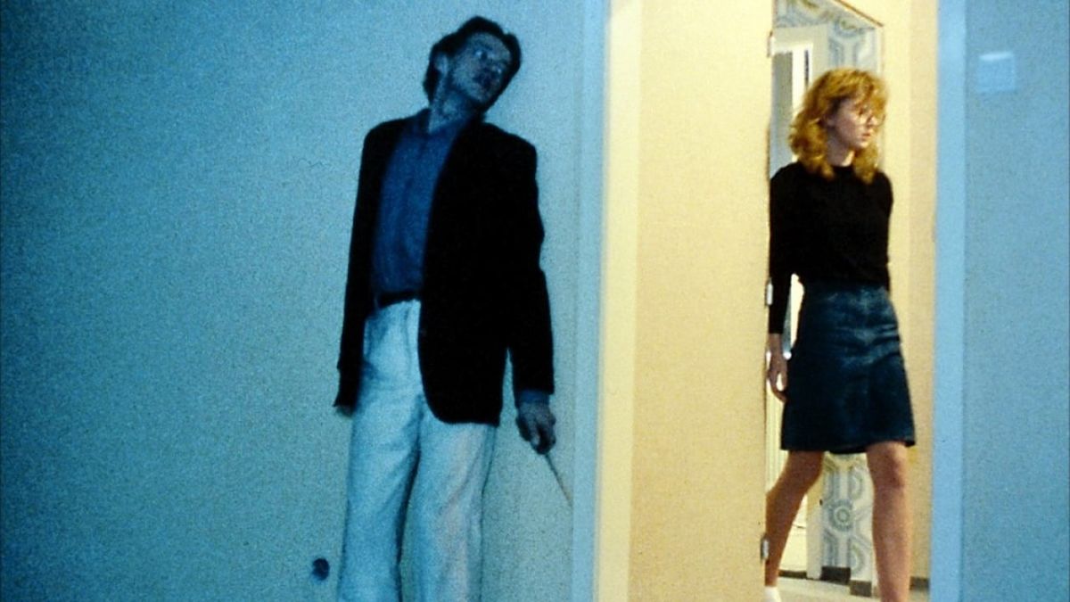 A dark-haired man in white pants, a blue button up and a black sport coat hides on the edge of a door frame, with a sharp object in his left hand. He looks up silently, waiting for his moment to attack. A blonde haired girl with large glasses, a black shirt, and a blue knee-length skirt walks into a lit hallway beside the man.