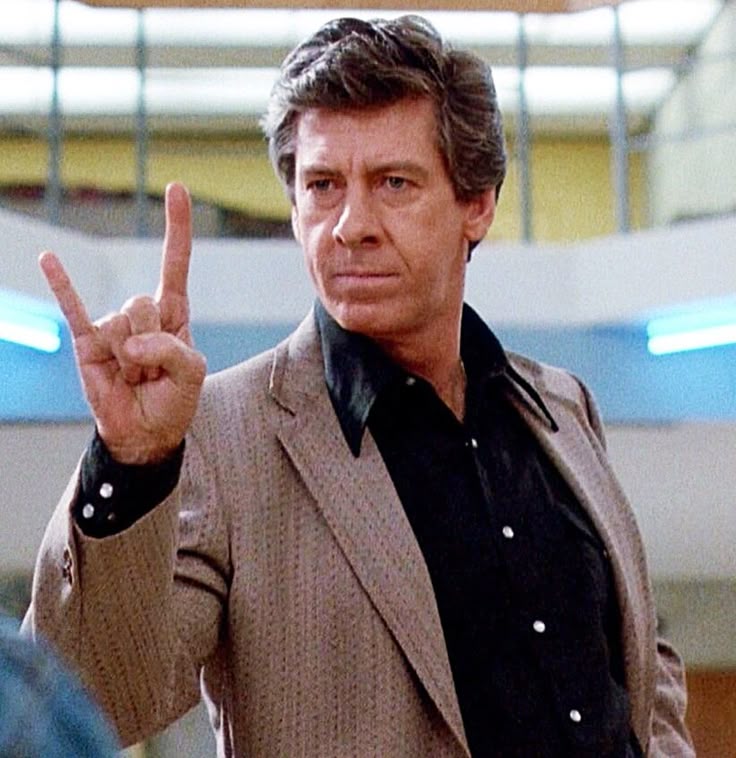 Paul Gleason, 40-year old man, in a 70s style brown suit stands with his right hand held up, index and pinky fingers extended to show two horns. His face is stern.
