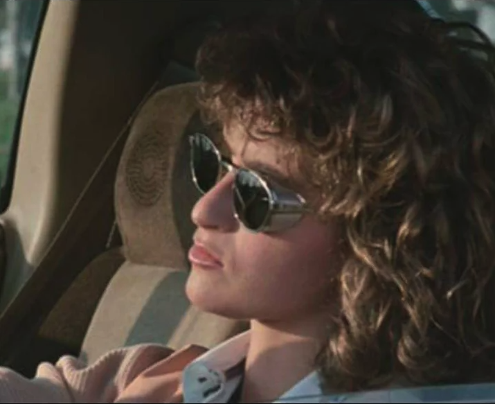 A young teenage girl with blonde fluffy hair, Jennifer Grey, sits n the driver's seat of an old car, with black, thin-framed sunglasses on. She looks forwards, distastefully, her eyes covered by the opaque glasses.