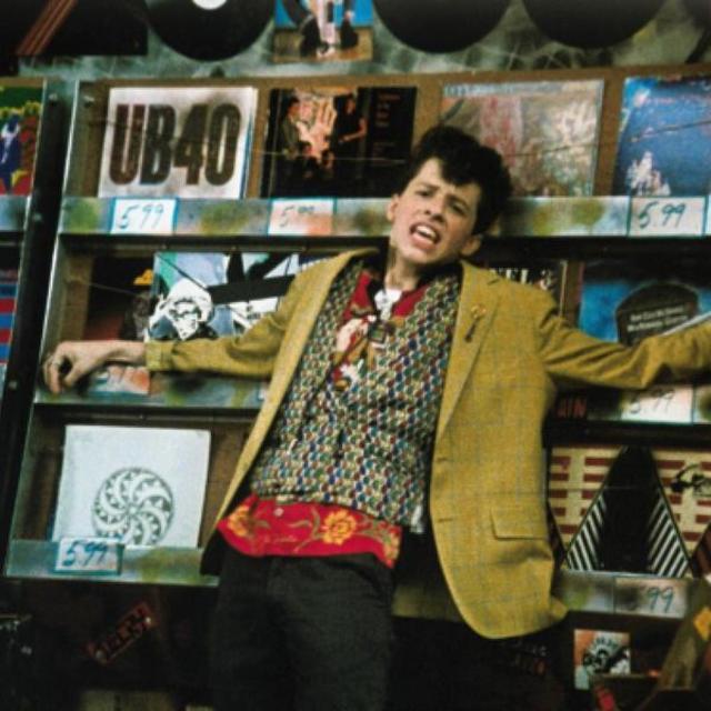 A young man with brown coiffed hair in an elclectic and colourful outfit leans against shelves of vinyl records. He is mid-sentence, his expression pained and yearnful.