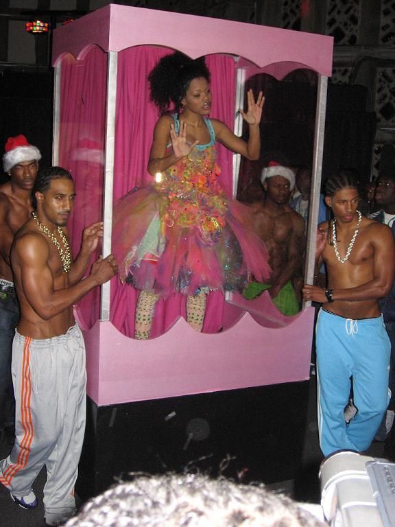 TV still from My Super Sweet 16. A teen girl stands in a huge Barbie doll box, wearing a colourful dress. The box is held by four shirtless men, some wearing chains, others wearing Santa hats.
