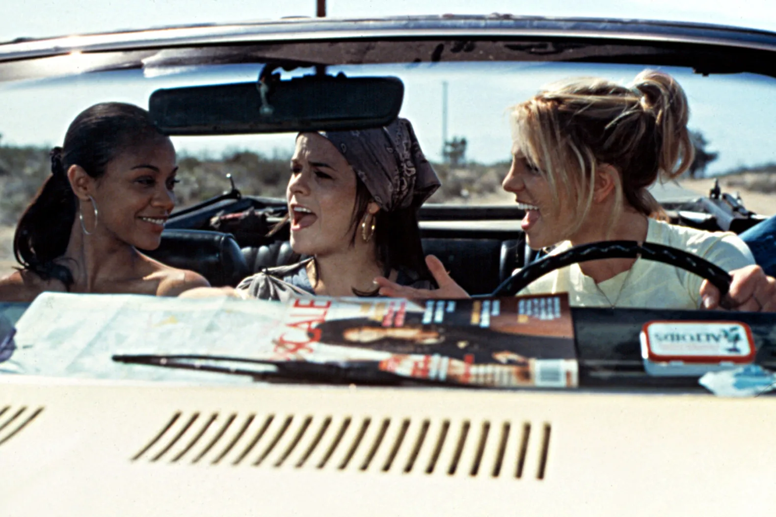 Three young women sit in the front of a convertible with the top down. On the left, a brown-skinned woman with her dark hair in a low ponytail and silver hoop earrings smiles at the other two. In the middle a fair-skinned girl with a purple bandana over her dark hair looks out left, her mouth wide open, as if singing. On the right, a blonde fair-skinned woman in a yellow shirt looks at the other two girls, her hair in a bun, and her mouth open, as if singing. The blonde-haired woman has her hand on the black steering wheel of the car. On the dashboard there is a map, a blurry magazine featuring a dark-haired, light-skinned model, and a tin of altoids, from left to right.
