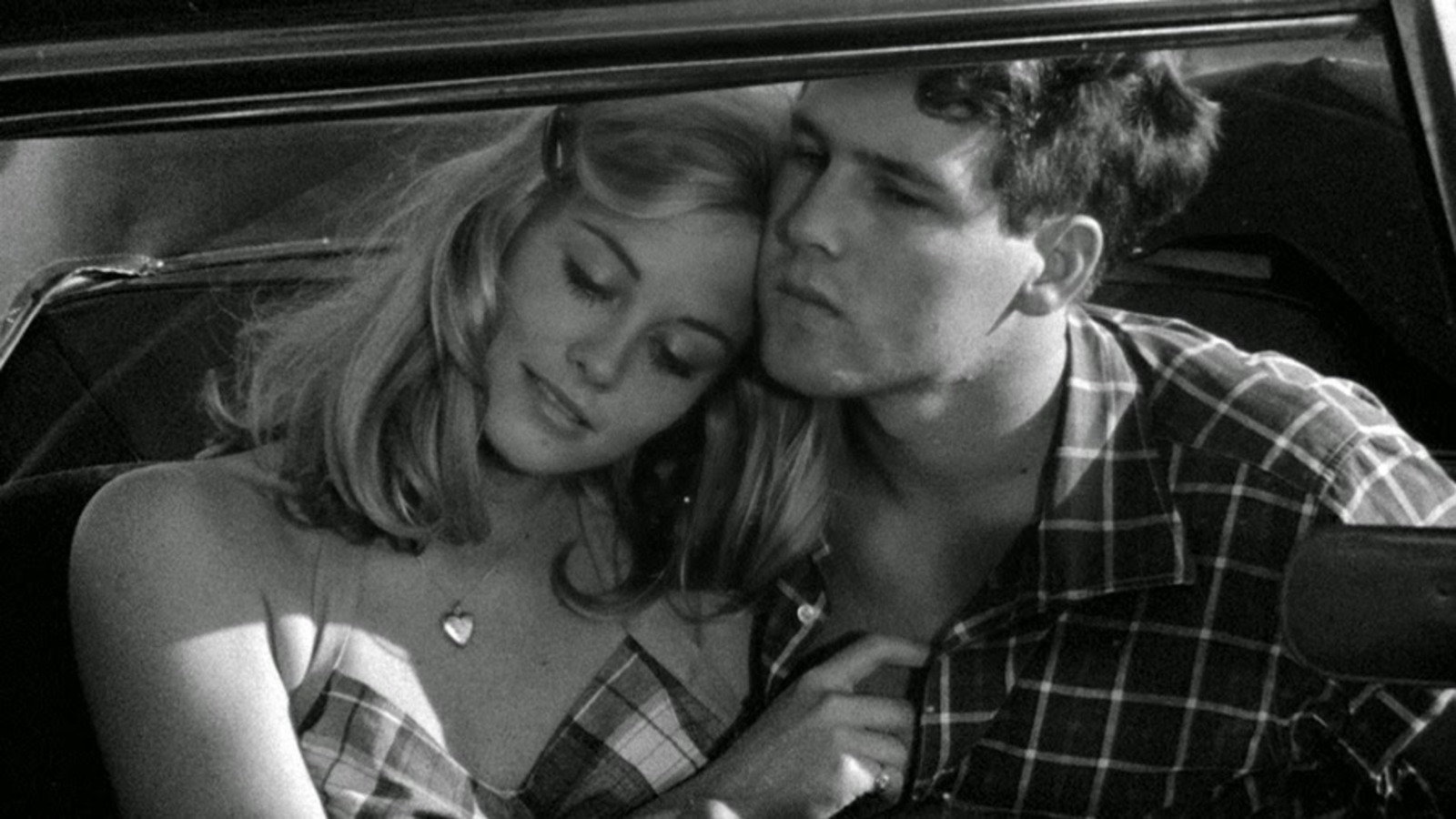 In black and white, a young boy and girl sit together inside a car. On the left, the blonde fair-skinned girl wears a gingham dress and a gold heart locket. She looks downward, her head tucked into the side of his neck while touching the boy's chest. On the right, the boy looks down, wearing a dark flannel shirt.
