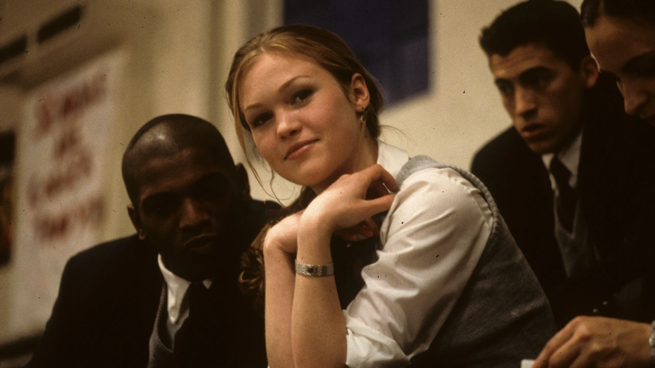 A fair-skinned blonde woman in a grey and white school uniform sits facing us and slightly smiling, her hands tucked just beneath her chin. Beside her on the left is a dark-skinned man in a similar school uniform sits facing us, his lips slightly puckered. Behind her is a fair-skinned dark haired boy in a similar uniform. They sit on the bleachers.