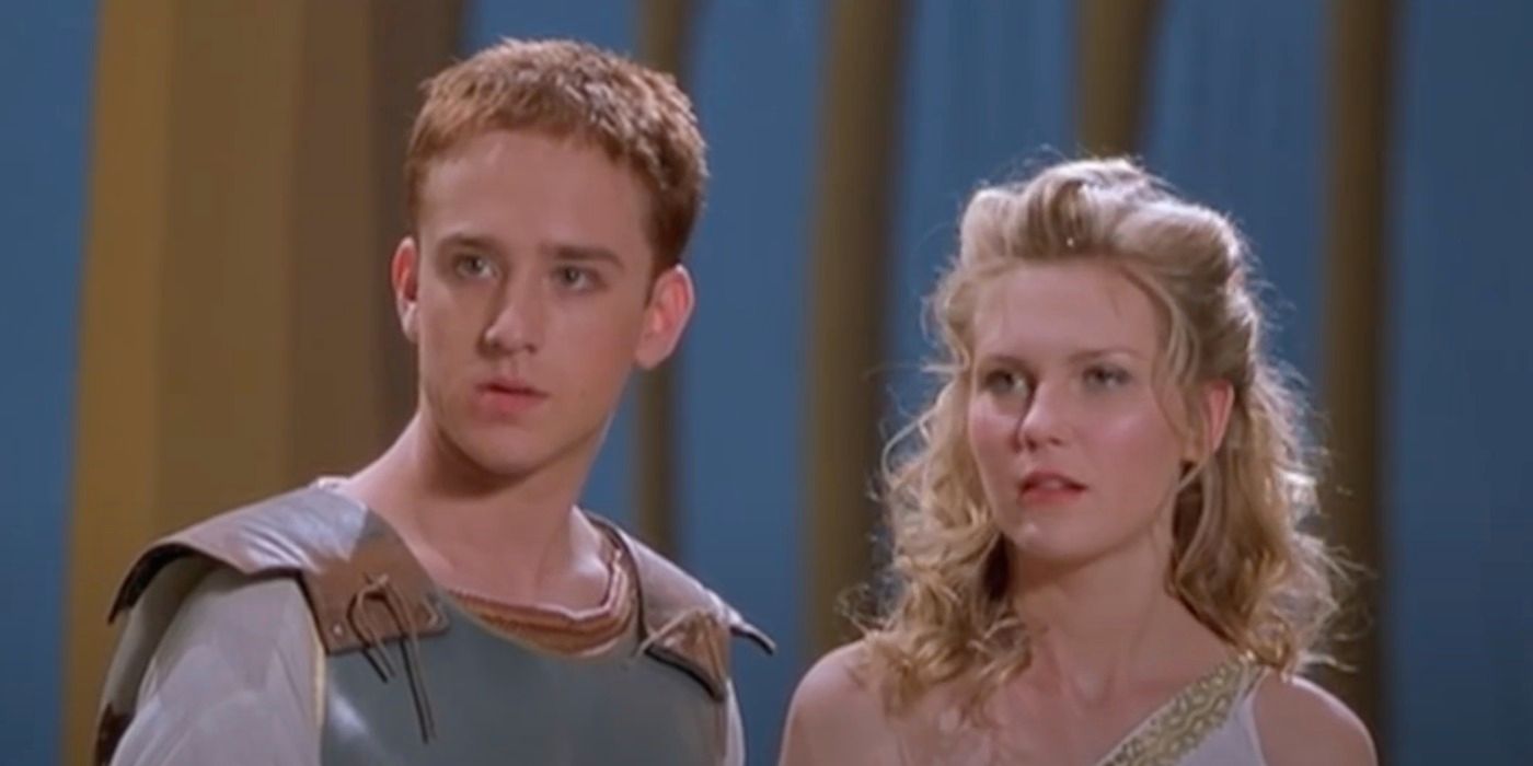 In front of a blue background with different width brown columns, a young fairskinned man with light hair looks to the left. He is dressed in leather armour. Beside him, to the right, a fair-skinned blonde haired woman in one-strap white garment looks forward to the left with him.