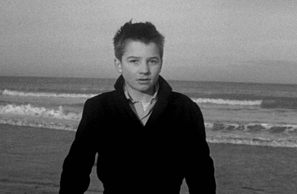 In black and white, a young man in a black jacket with short hair stands in front of a beach shore, his face neutral, almost caught off guard.