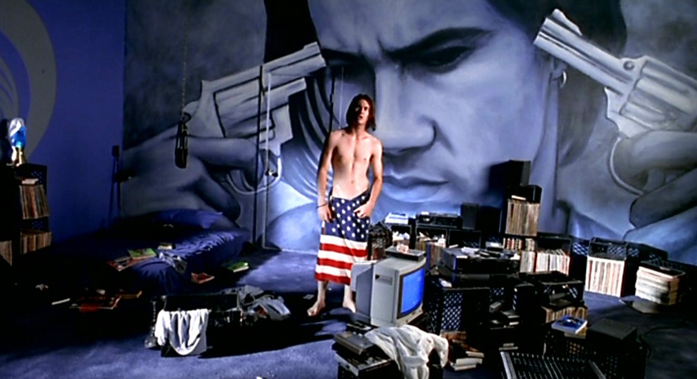 In a large dark room, a young man stands in the middle, behind a large black computer desk, his lower half covered by an American flag towel. In the left corner, a blue bed with no bedframe, and a large mural of the young man with two revolvers pointed by himself at his own head on the back wall.